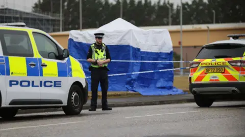 A police officer standing in front of a police cordon and blue forensic tent and two police vehicles in Arbroath.