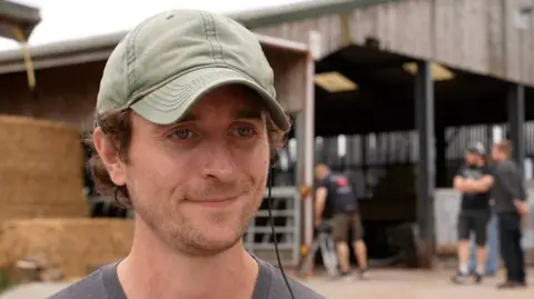Alex Challies, First Assistant Director of MacDonald is wearing a grey t-shirt and green baseball hat. He has a black wired headphone in his left ear. In the background are more crew members from the shoot and a bale of hay.