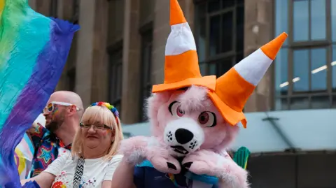 A Pride parade participant wearing a large pink animal costume head topped with two orange-and-white traffic cones poses beside other marchers carrying colourful flags in a city street parade.