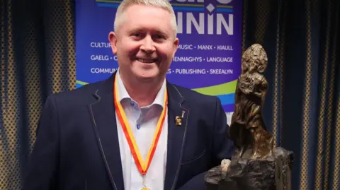 Juan Garrett, who has short grey hair, is smiling. He is wearing a blue blazer and has the yellow and red ribbon of the RBV medal around his neck, and is standing next to the trophy, which is a bronze depiction of Manannan.