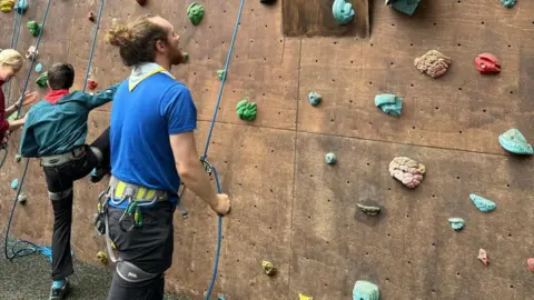 A man looking up at a climbing wall wearing a blue scouts top and neck scarf, with a young boy about to climb the wall next to him talking to a different man.