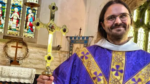 Rev Robin Thwaites the vicar of St Breward Church on Bodmin Moor in his robes cluthing a brass cross with inlaid stones standing in front of the altar