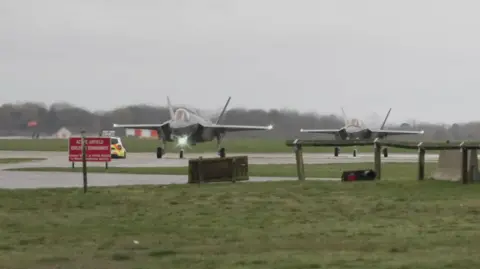 Two planes, which appear to be F-35As from the Joint Strike Fighter programme, located at RAF Lakenheath. They are located on a runway or taxiway, surrounded by grass lawns. A police car is visible. 