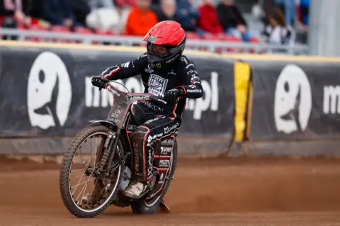 TŻ Ostrovia/Wojciech Tarchalski A motorcycle speedway rider in a black race suit and a red helmet leans into a turn. A trail of dust is behind his bike. Spectators and advertising boards are in the background.
