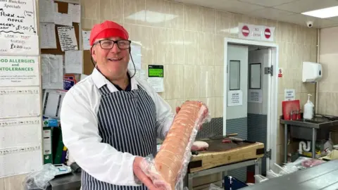 Andrew Turner/BBC Jim Wyllie is wearing a butcher's white coat and blue and white striped apron, and a red flat cap. He is holding a joint of rolled stuffed pork belly. The back wall is tiled, and covered in notices and orders. There is a butcher's block and stainless steel sink and worktop.