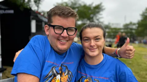 A man wearing thickly rimmed black glasses sits next to his friend, Both are wearing matching blue T-shirts and smiling. He gives a thumbs up. 