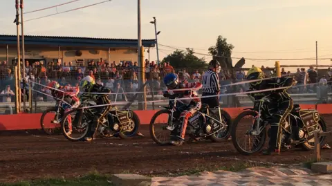 Riders are getting ready behind the starting line at Swindon Speedway. People are watching from the stands.