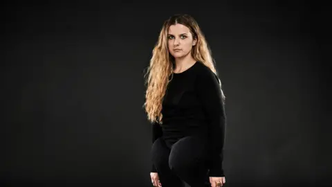 David Myers Ella Glendining wearing a black shirt and jeans stands and looks at the camera. She has long blonde hair. Ella has a congenital, rare leg disability.