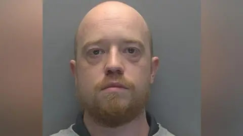 Liam Brodie is seen in a police custody shot. He is looking at the camera and has a balding head with a ginger beard and moustache. He is wearing a grey sweatshirt.