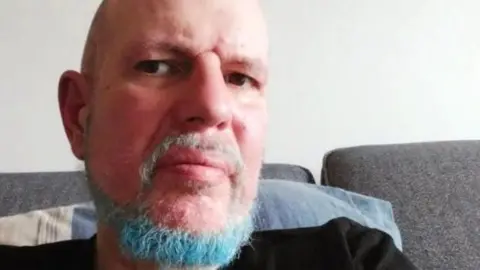 A bald man with a blue and grey coloured beard and wearing a black top, takes a selfie while sitting on a grey sofa, with a blue and white cushion behind him.