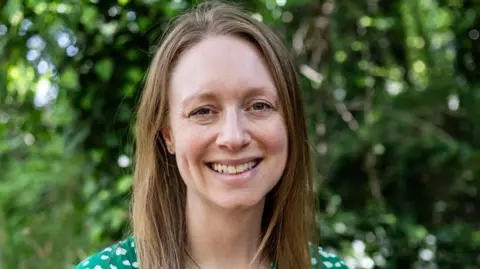 Supplied A headshot style photo of Rachel. She's smiling at the camera. She has shoulder length brown hair and wearing a green dress. Behind her is trees and bushes.