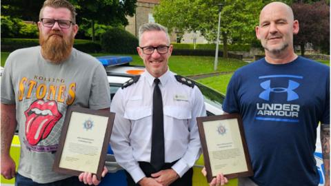 Three men - two holding certificates with a police officer in the middle 
