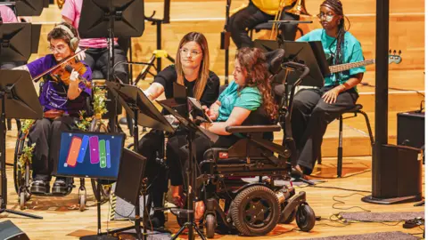 National Open Youth Orchestra Evie Read is sitting among other members of the orchestra on a stage reading sheet music while using a tablet.