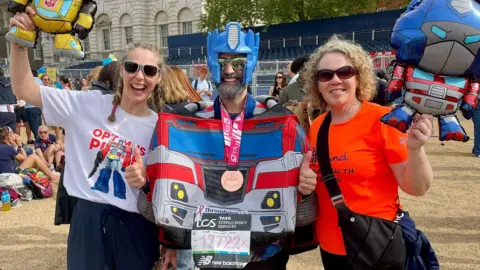 Matt Batchelor Matt Batchelor and two women, pose together after the London Marathon. Batchelor is wearing a large blue and red Optimus Prime costume, with only the head visible. His head is covered in a blue plastic helmet, and he is wearing reflective sunglasses. He has a pink race medal, and the two women beside him hold robot inflatables.