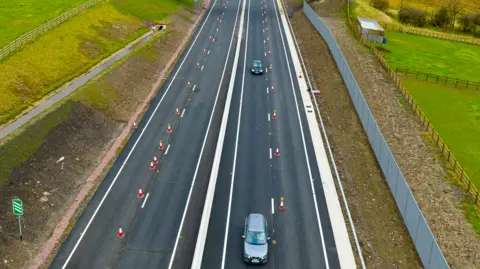 Cars pass along the Heads of the Valley road, with two lanes going in each direction. A row of cones runs alongside the middle of the lanes on each side. Grass verges can be seen on either side. 