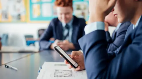 Young male student in a blue school uniform is pictured checking his phone in a classroom.