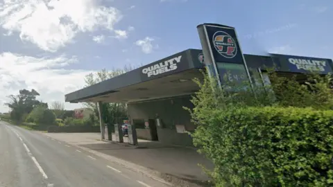 Google A view looking south on Lancaster Road in Cockerham. On the right a garage forecourt can be seen with black and white signs reading 'quality fuels'. On the left of the single carriageway road is a green hedge.