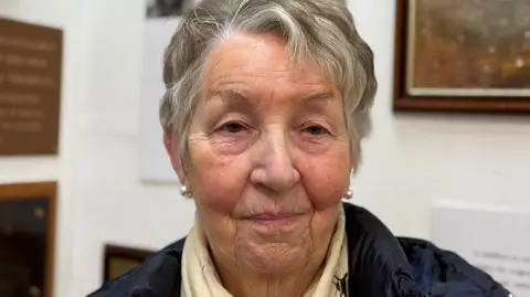 Head and shoulders image of Ann Parkinson standing in front of the exhibition. She is wearing a navy coat and cream scarf, has short wavy grey hair and is smiling