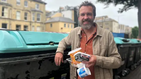 a bearded man stands next to a line of green recycling bins. He is wearing a beige linen jacket over an orange linen shirt and is holding four empty plastic tubs. He is smiling at the camera, with his elbow casually rested on one of the bins