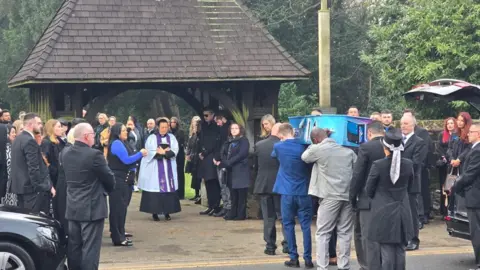 Mourners gather outside a church for the funeral of Leo Ross. A group of men carry Leo's coffin as they walk towards the church. The coffin has been painted with artwork - it is blue and has items, such as games consoles, on the sides. A vicar - a woman with black hair holding a bible to her chest - stands in the middle of a path, with mourners on either side.