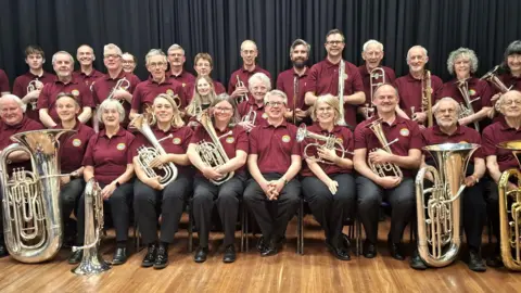 A brass band, all dressed in maroon shirts and black trousers, is sitting down for a photograph. Some of them are holding instruments.