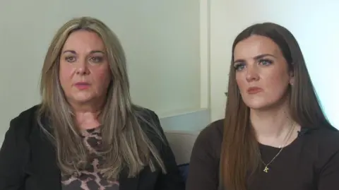Joanne and Kelly sit together as they do a tv interview. Joanne has long blonde hair and weara a dark jacket over her top. Kelly wears a dark top.