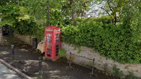 Google A red phone box on a pavement. There is a brick wall behind it with green bushes and trees