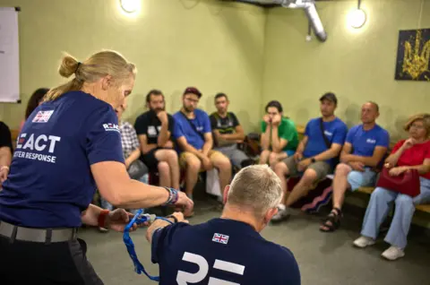 Lewis Inman A group of people in blue, red and green t-shirts are sitting in a row of chairs around the edge of a room. In front of them, two people, wearing navy t-shirts with the union jack on are demonstrating how to operate a tourniquet.