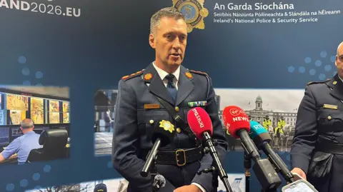 Garda officer wearing a blue uniform. He has short dark and grey hair and has mics in front of him. An Garda Siochana is on the background behind with police pictures.