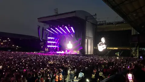 Jon Wright/BBC Ed Sheeran performs on stage in Portman Road. It is dark and the stage is lit up. Thousands of fans watch on.