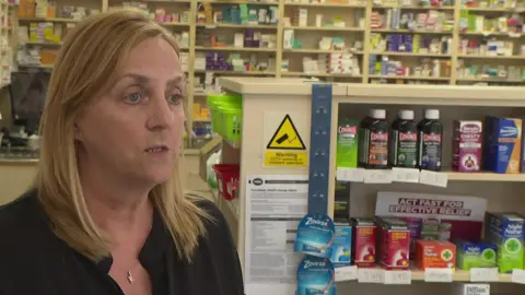 Pharmacy manager Sue Jayne with long blond hair wearing a black tunic. She stands in a pharmacy with medication and tablets on shelves behind her.