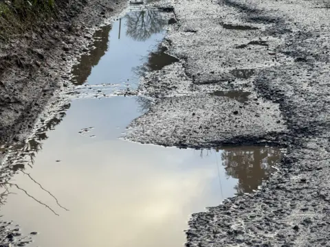 A damaged section of tarmac at the edge of a road. Different layers of road surface are visible to the right. On the left of frame a deeper hole is full of water. Reflections of trees and hedges can be seen.