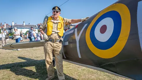 Andy Bell Images A man in vintage RAF-style uniform with sunglasses and a pipe in his mouth stands beside the wing of a replica World War Two aircraft on a grassy field. The aircraft’s large RAF roundel is clearly visible on the wing, while visitors and seafront buildings can be seen in the background under a clear blue sky.