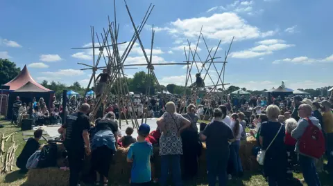 Natalie Malcolm/BBC Crowds gather round a small outdoor stage with bamboo poles, and two performers doing something on the stage. It is in a park with trees on the far side, and is a sunny day. Small kiosks and tents line the other side of the stage.