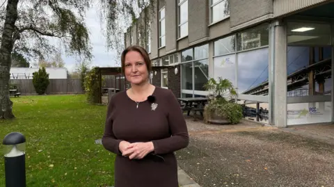 A woman wearing a brown dress stands outside a grey building. A green area of grass is next to her. The building has many large windows. The woman is smiling at the camera and is holding her hands in front of her. 