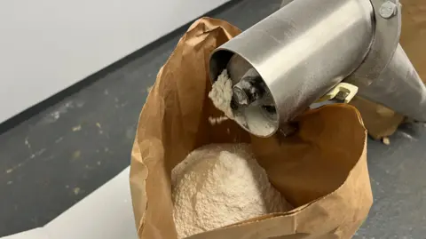 John Devine/BBC A steel tube has white flour coming out of it into a brown paper sack.