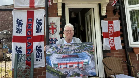 BBC Mary, a woman with short white hair and glasses, stands holding a print of Old Trafford in front of a house covered with various England flags.