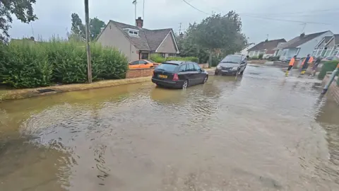 A burst water main in a road, showing a large amount of water. There are two people in high vis outfits in the distance, and houses too. Two cars are parked on the street. 