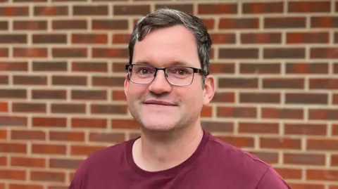 Peter Smith is wearing a red T-shirt and has short, dark hair.