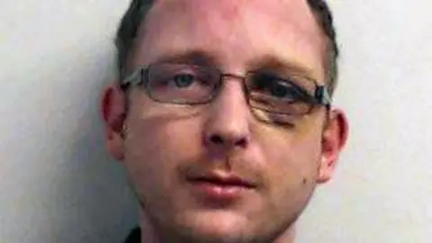 Man with short hair and glasses looking at the camera against a plain light-coloured wall.