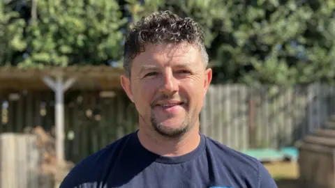 A man is smiling at the camera with short curly black hair, a moustache and beard. He is wearing a blue t-shirt
