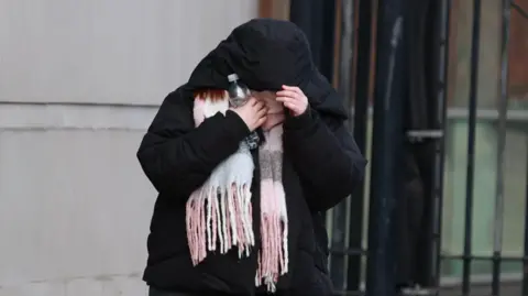 Liam McBurney/PA Wire Laura Graham pictured outside the court in Belfast.  She is wearing a black overcoat, the hood of which she is using to cover her face.  She is also wearing a fringed pink. gray and white scarf and is holding a plastic bottle.  