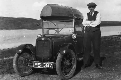 Shetland Museum and Archives Photographer John D Ratter standing next to an old-fashioned black car that has a small boat strapped to the top. He is well dressed with suit trousers, a vest and shirt. He is wearing a flat cap. 