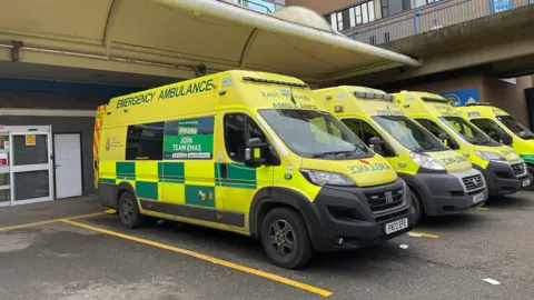 A row of yellow ambulances parked outside QMC under a white roof