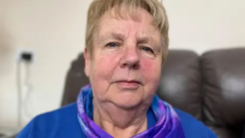 Helen Fraser looks directly at the camera. She is sitting in a leather armchair. She is wearing a blue jumper and neck-scarf with a fancy pattern