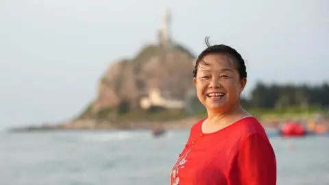 Su Min A smiling Su Min in a red top standing by a water body with a blurred hill in the background. 