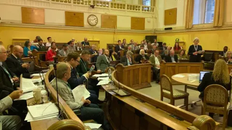 BBC A council meeting is taking place. A woman is standing and speaking.