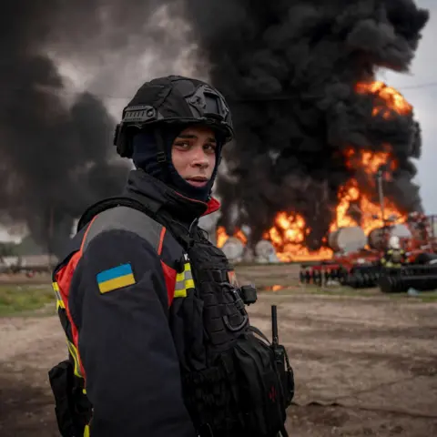 Valeria Demenko/DSNS members of emergency services in the northeast of East Sumy, dressed in a high Vis, with the Ukrainian flag, shaded on the hand and combat helmet and balaclava, watches fatigue, while standing in front of a burning gas warehouse near the Russian border.