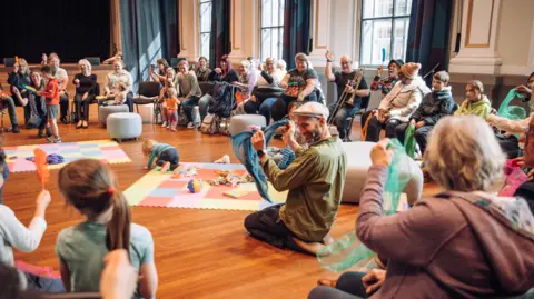 Bristol Beacon Lots of people sitting in a circle in a workshop at the Bristol Beacon. There are soft mats in the centre with children playing, people waving silk scarves and others with instruments. 