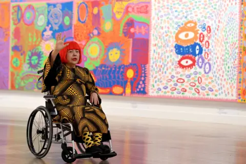 Getty Images Kusama in a wheelchair with red hair and black and yellow spotty clothes, in front of large multi-coloured paintings